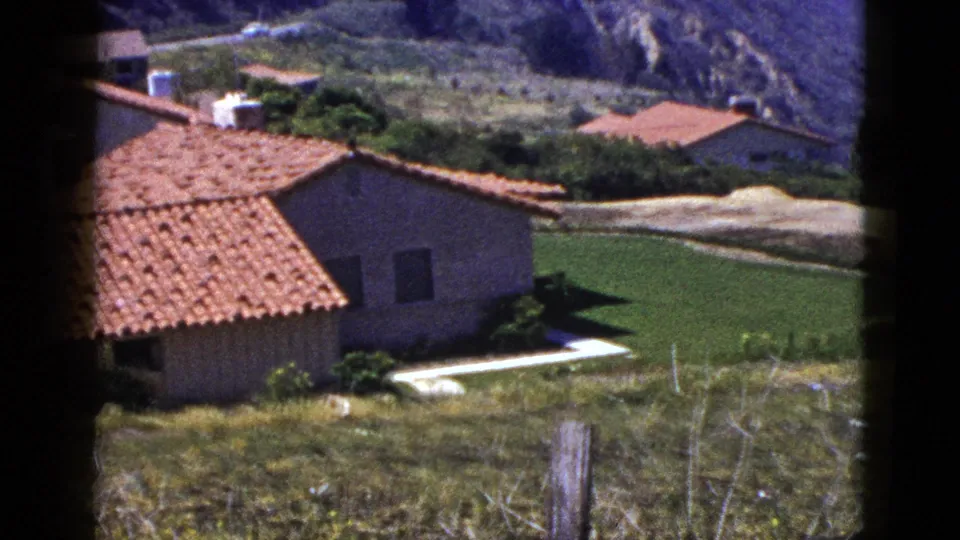 1953 Palm Springs frame showing a red tile roof house beside a green lawn, with a pale hillside road and mountains beyond.