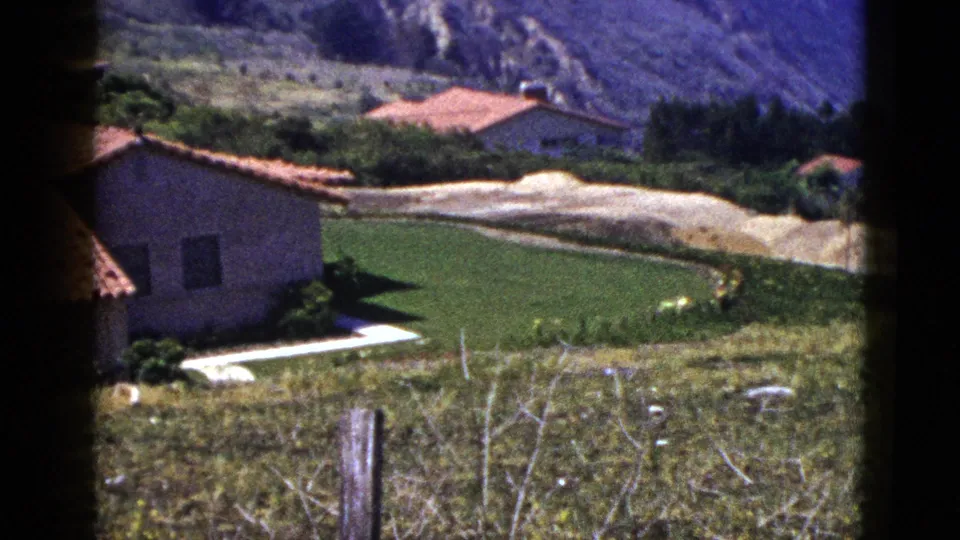 Palm Springs residential hillside view with a stucco house, terracotta roof, lawn, pale drive or road, fence posts, and distant mountains.