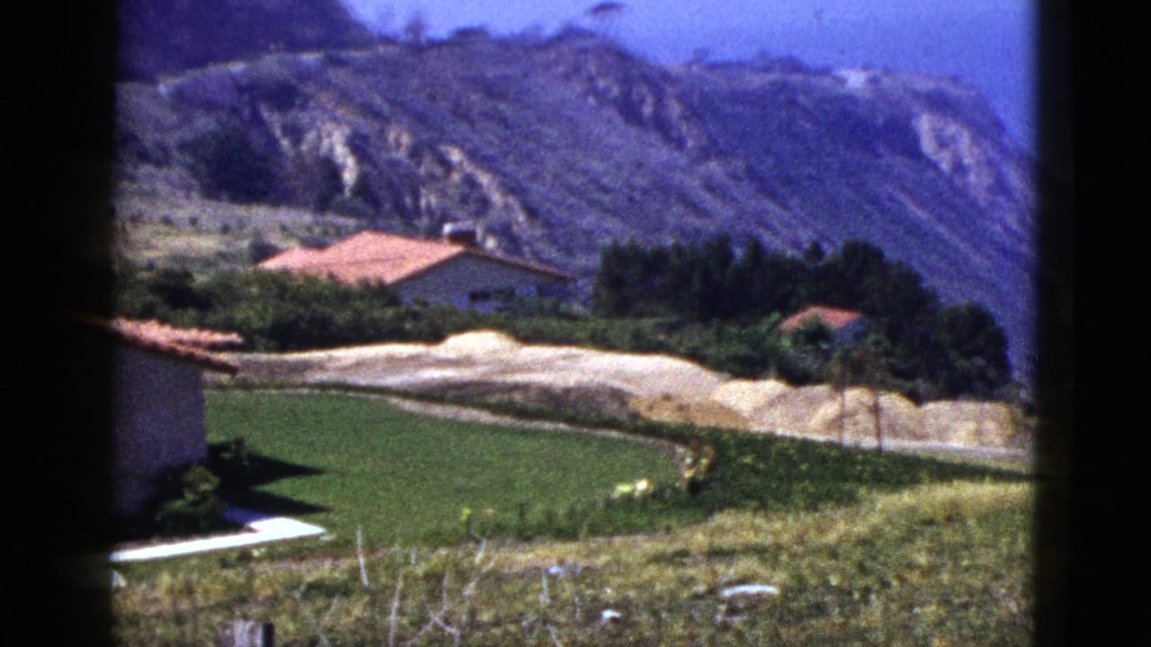 Red-roofed houses, lawn, pale hillside, and mountains.