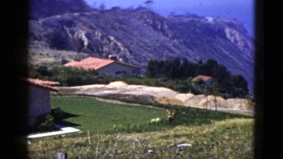 1950s 8mm frame showing red-roofed hillside houses, a green lawn, pale graded earth, and rocky slopes in the distance.