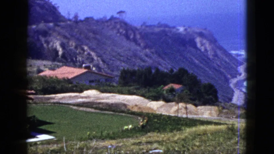 1950s 8mm coastal view with hillside homes, green lawn, pale earth, steep cliffs, and ocean surf beyond.