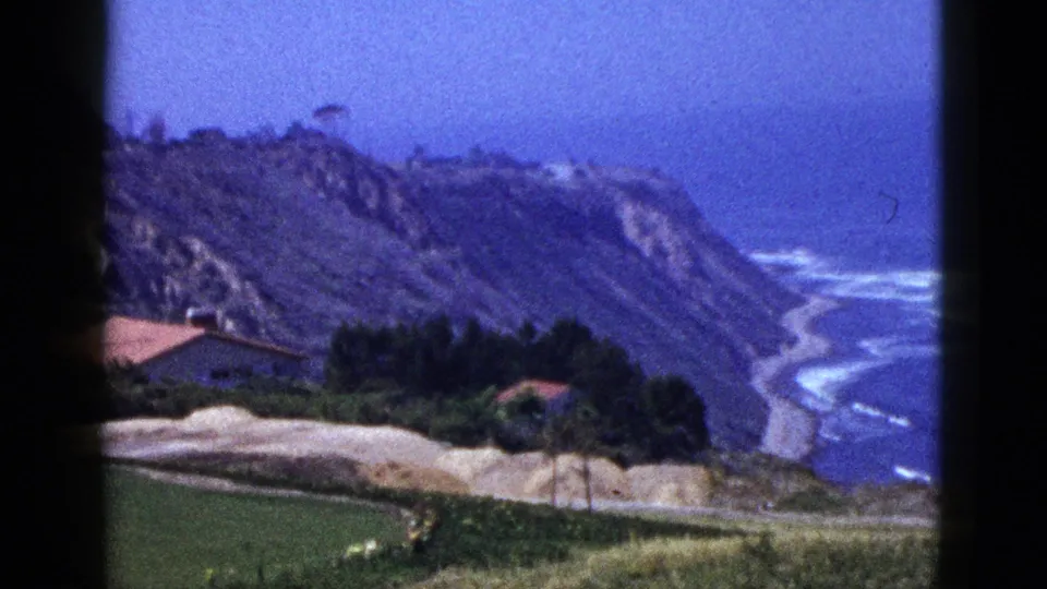 1950s 8mm frame of a coastal hillside with red-roofed homes, green lawn, cliffs, and Pacific surf in the distance.