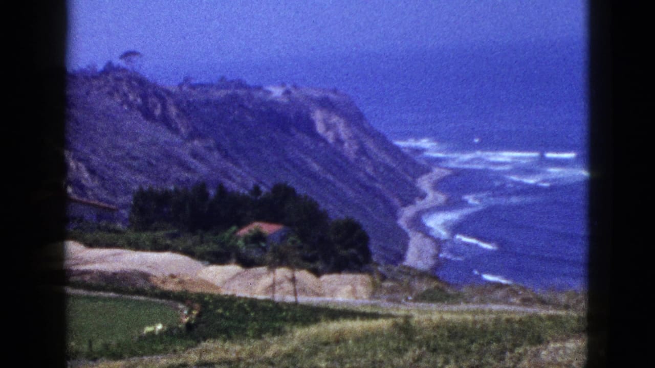 1950s 8mm home-movie frame of a California coastal overlook with cliffs, surf lines, grassy hillside, and a small red-roofed building.