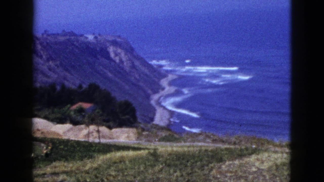 High view of cliffs, ocean waves, hillside grass, trees, and a small red-roofed building.