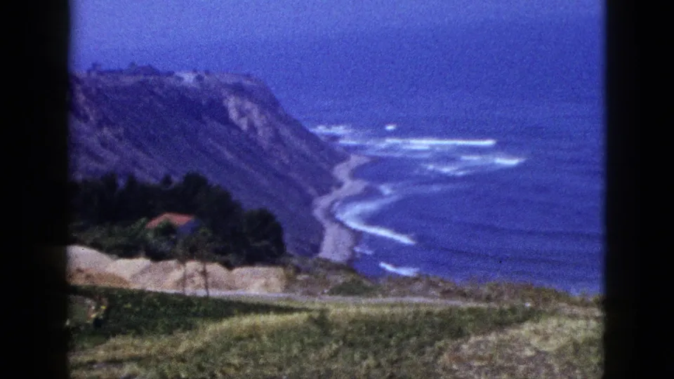 1950s 8mm frame looking over a California shoreline, with steep cliffs, blue water, white waves, trees, and a red-roofed structure below.