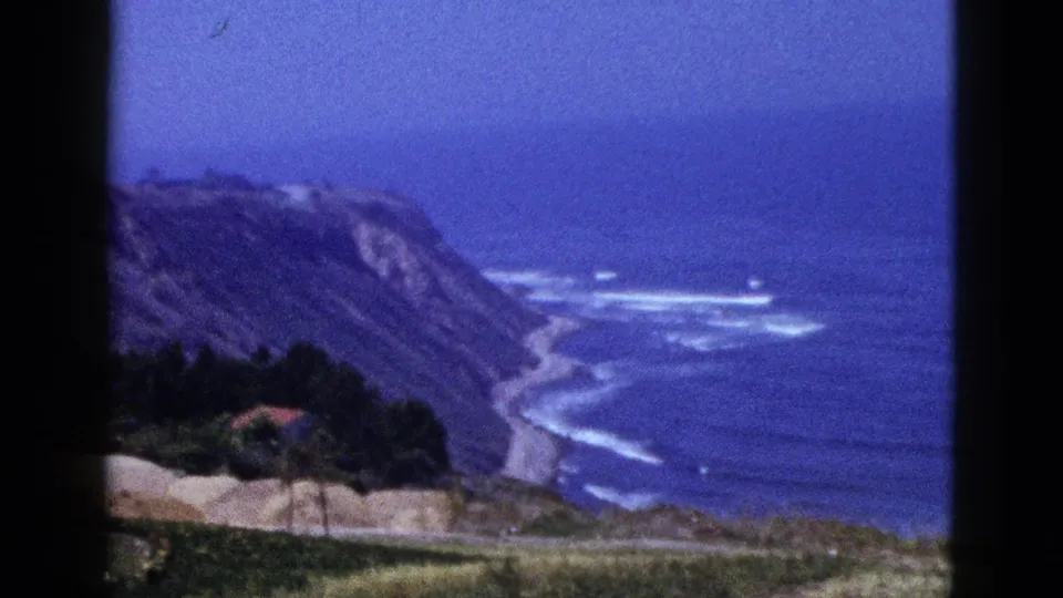 1950s 8mm coastal frame showing a cliff-backed shore, blue ocean surf, hillside vegetation, and a small red-roofed structure near trees.