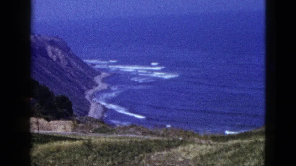1950s 8mm home-movie frame of a California coast view, with blue ocean, white surf, cliff slopes, road or shoreline curve, and grassy foreground.