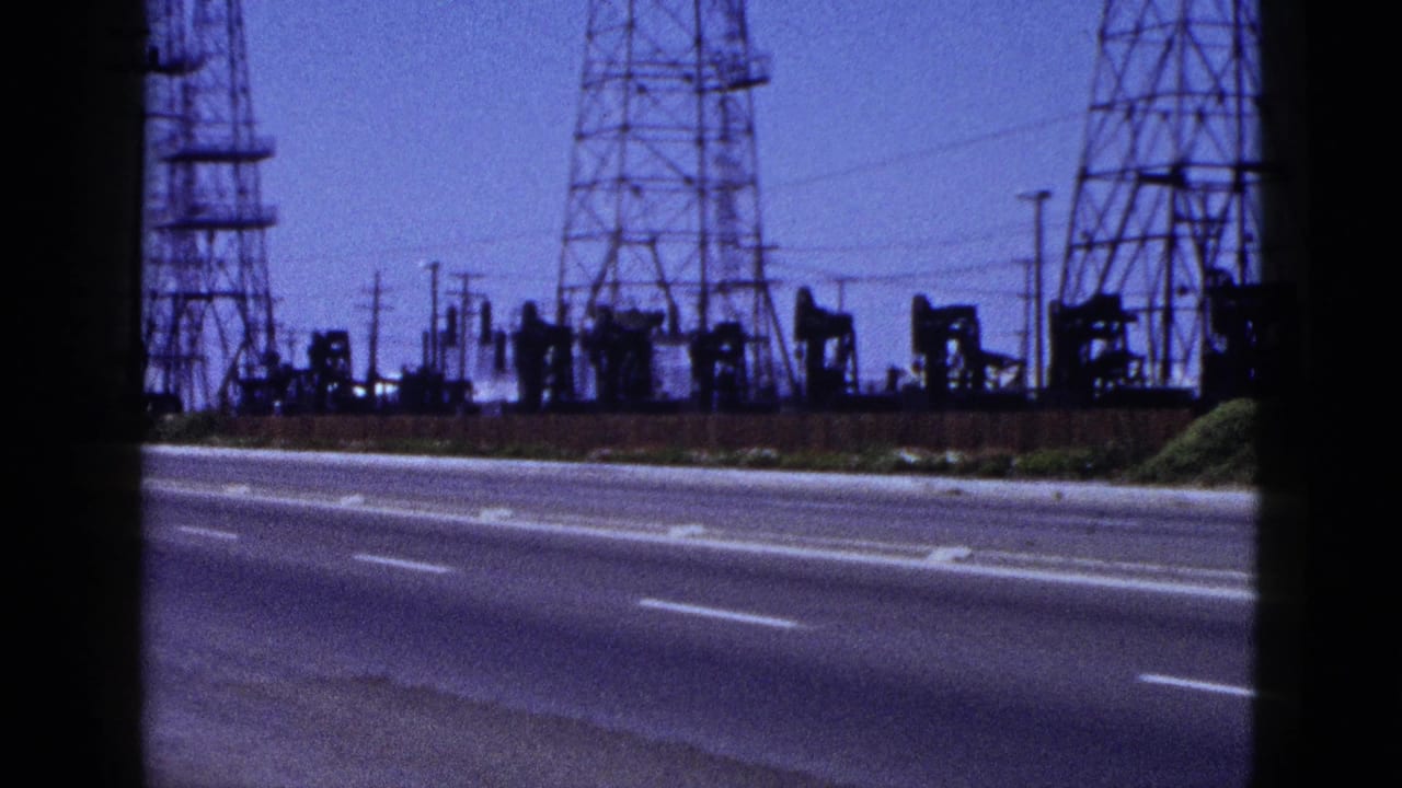 Metadata-dated 1953 8mm frame shows a California roadside with lattice power towers, utility wires, and oil pumpjacks in the distance.