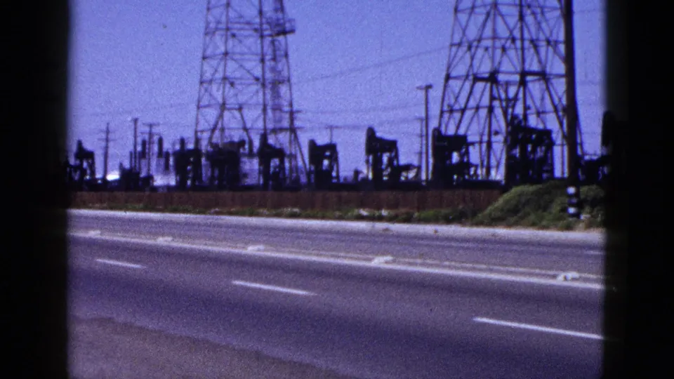 A 1953 roadside 8mm frame shows pumpjacks, lattice transmission towers, poles, and white lane markings under a blue cast.