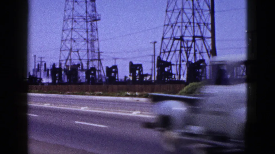 A passing vehicle blurs across a 1953 8mm roadside view of power towers, utility wires, and oil pumpjacks.