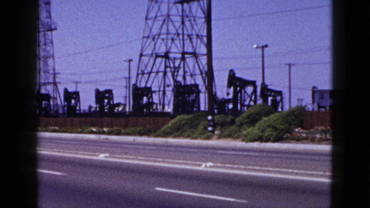 Roadside oil field with pumpjacks, towers, utility poles, and shrubs.