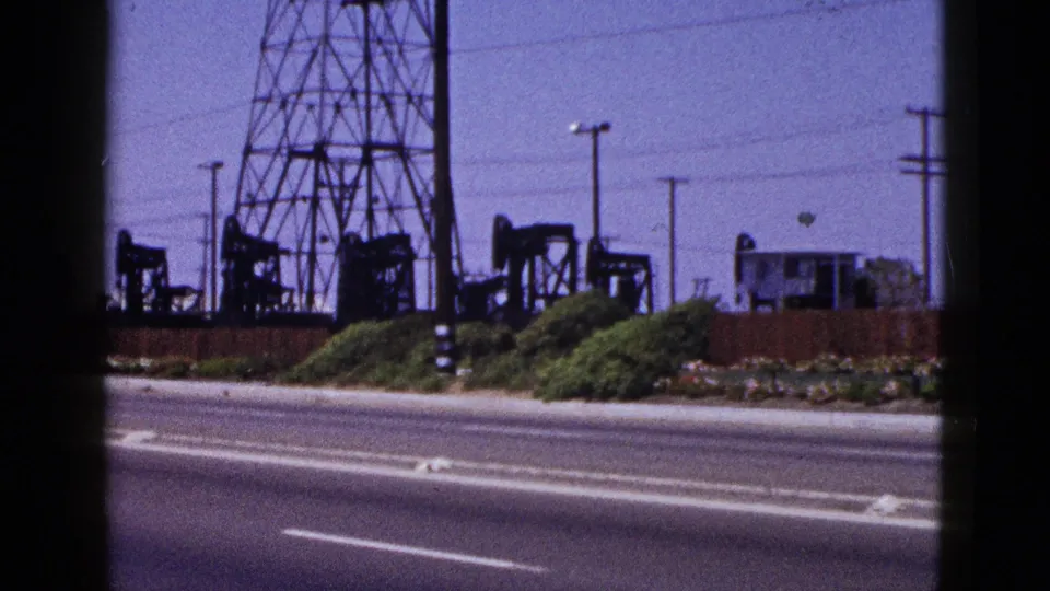 Roadside view of pumpjacks, transmission tower legs, shrubs, utility poles, and a small structure in Palm Springs.