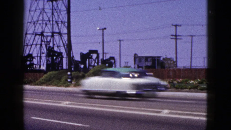 A white car blurs across the 1953 Palm Springs road view with pumpjacks, towers, shrubs, and utility poles behind it.