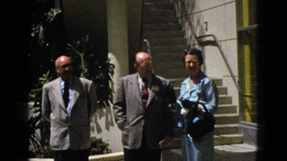 Three adults in dress clothing stand outside a Palm Springs building entrance, with steps, columns, plants, and a doorway visible nearby.