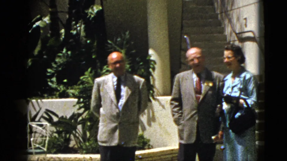 A home-movie view of three adults outside a Palm Springs building, including two suited men and a woman near steps and shaded landscaping.