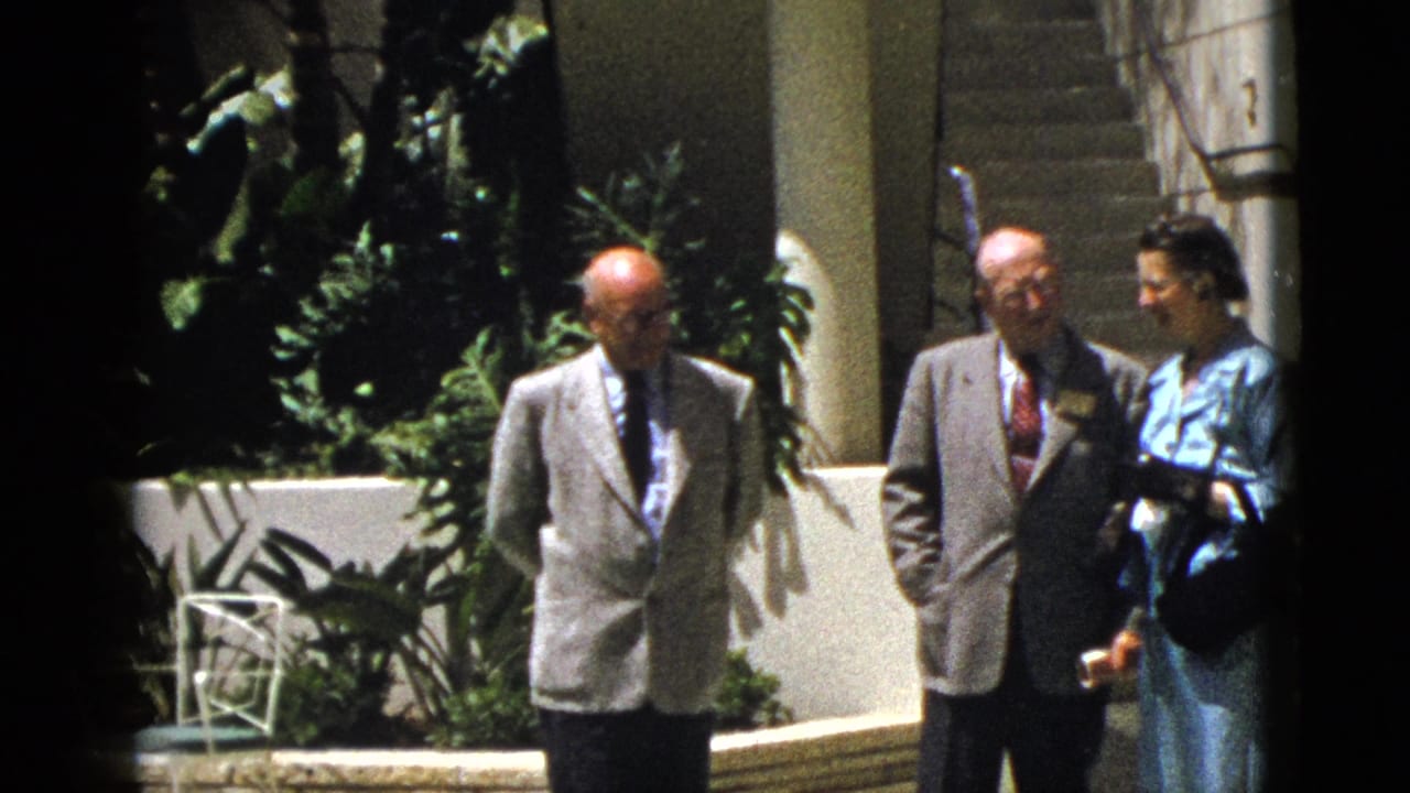Three adults stand outside near stairs, plants, columns, and a white wall.