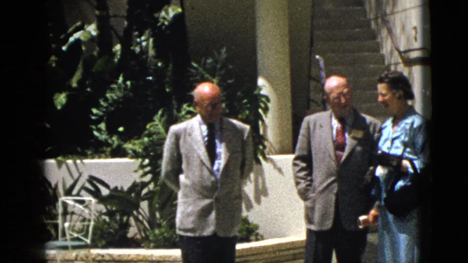 In 1953 Palm Springs home-movie footage, two men in suits and a woman in blue stand outside near stairs and greenery.