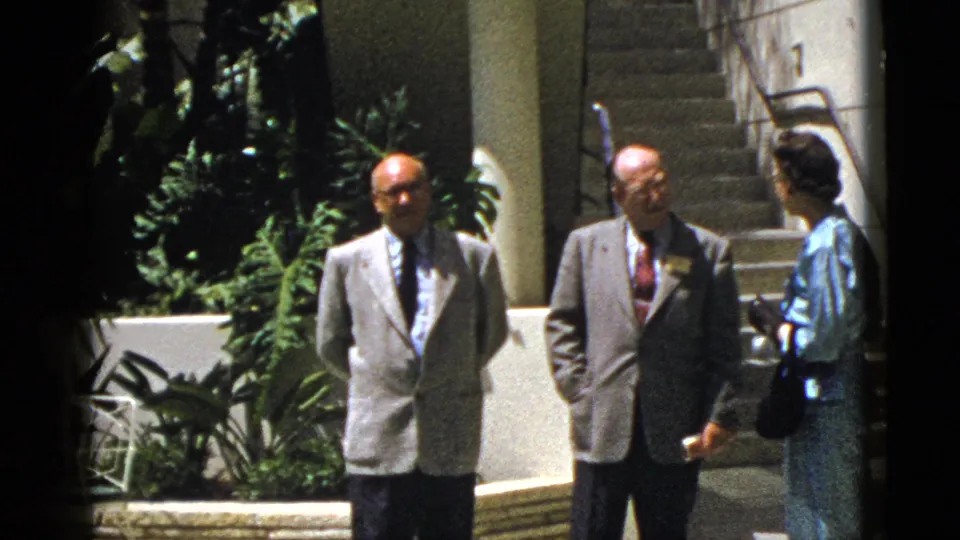 Two suited men and a woman in a blue dress appear to converse outside a Palm Springs building in 1953 footage.