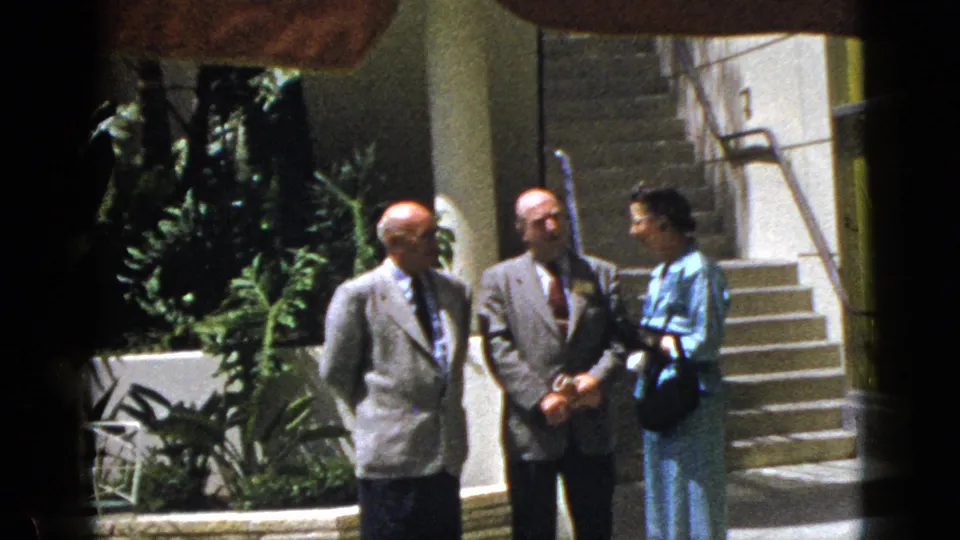 Three adults in mid-century dress continue their quiet outdoor conversation in Palm Springs, California, in 1953 footage.