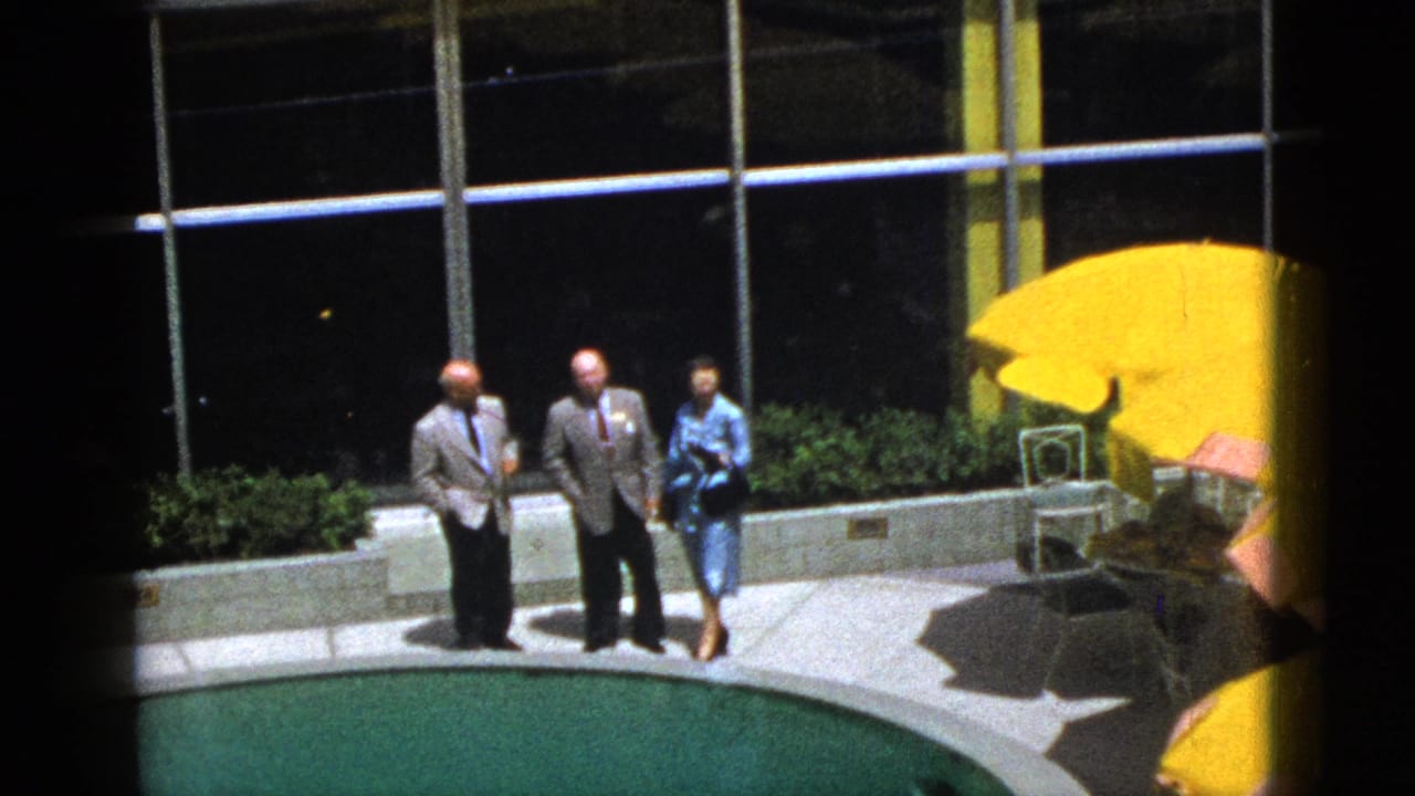 Two men in suits and a woman in a blue dress stand beside a swimming pool near yellow umbrellas.