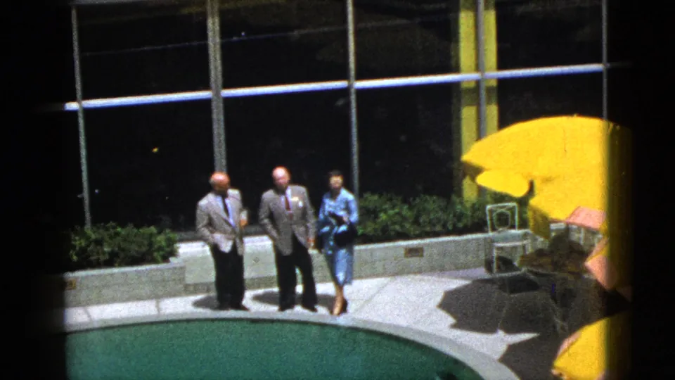1953 Palm Springs poolside scene with two suited men and a woman in a blue dress near glass windows, planters, and yellow patio umbrellas.