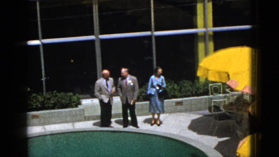 1953 Palm Springs poolside view showing two men in suits and a woman in a blue dress near patio furniture and reflective glass windows.