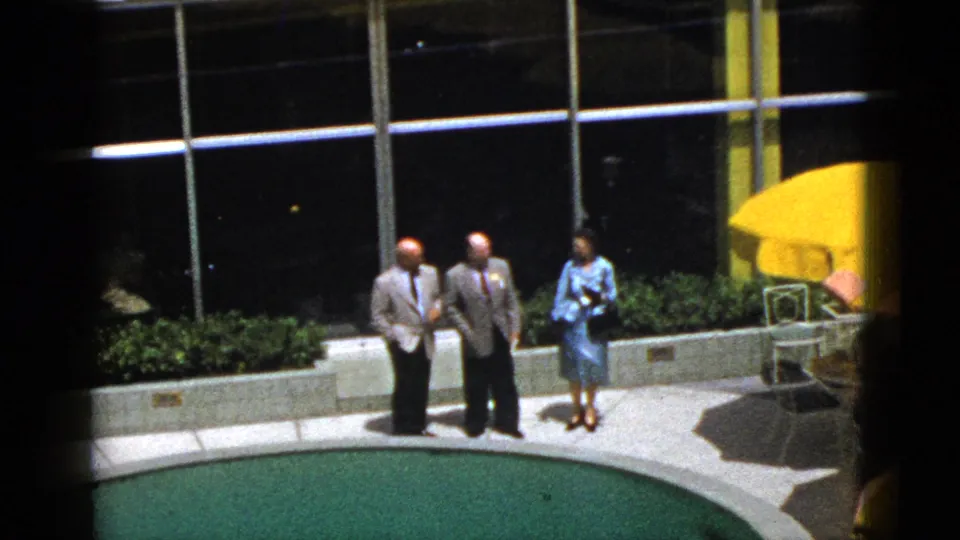 1953 Palm Springs home-movie frame of two suited men and a woman in blue beside a curved pool edge, planters, windows, and yellow umbrellas.