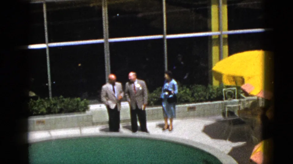 1953 Palm Springs poolside frame with two suited men and a woman in a blue dress near umbrellas, chairs, greenery, and a modern glass facade.