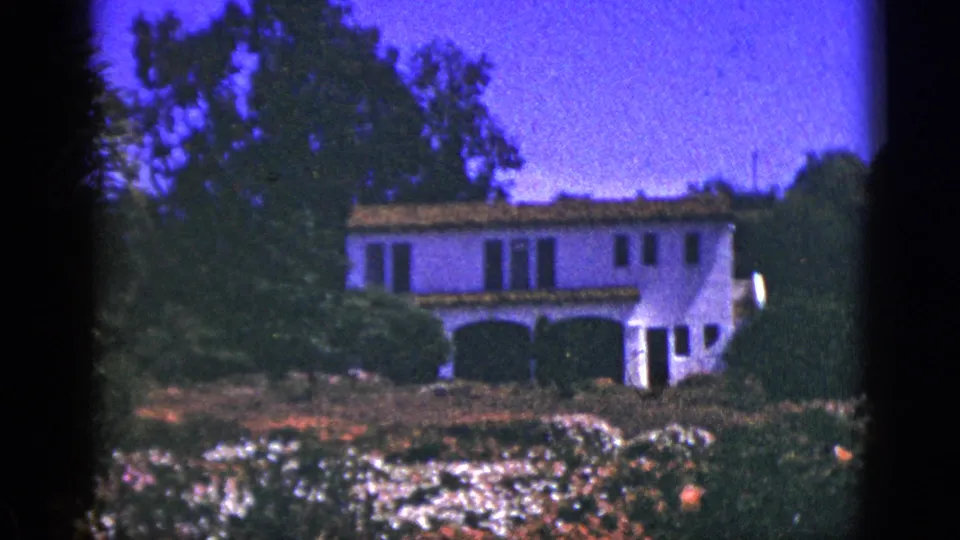 1953 Palm Springs home-movie view centering a white stucco house with tile roof, arched openings, flower beds, and tree cover.