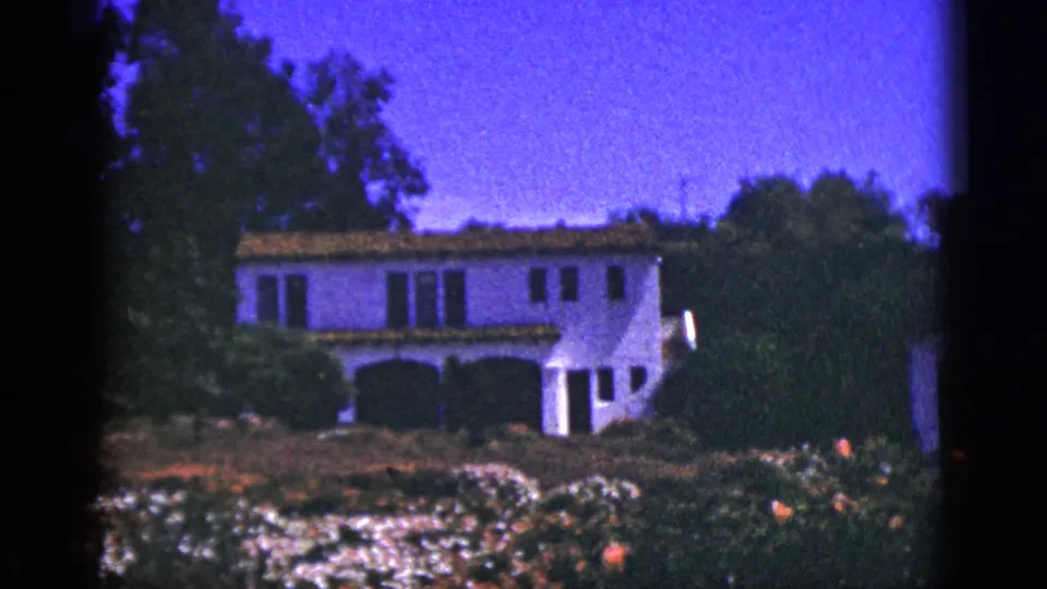 1953 Palm Springs frame showing the white stucco building, tile roof, arched porch, dense garden plantings, and dark 8mm frame edges.
