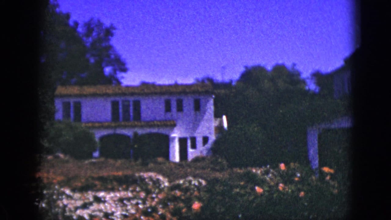 1953 Palm Springs home-movie frame showing a white stucco Spanish-style house behind a landscaped flower garden.