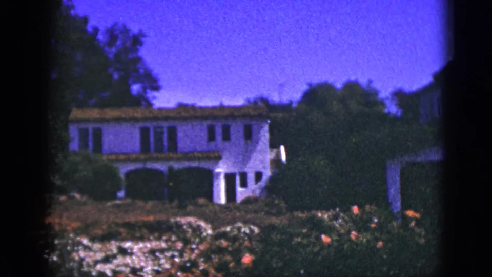 1953 Palm Springs home-movie frame showing a white stucco Spanish-style house behind a landscaped flower garden.