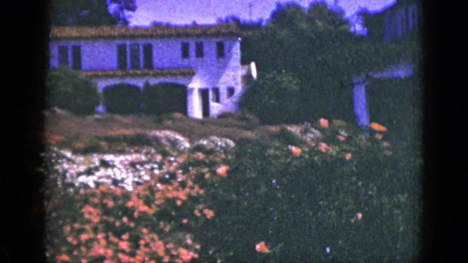 1950s 8mm frame of a white Spanish-style residence partly screened by shrubs and blooming garden plants.