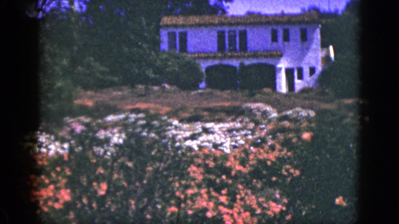 1953 Palm Springs home-movie frame showing a white stucco house with terracotta roof partly obscured by flowering garden plants.
