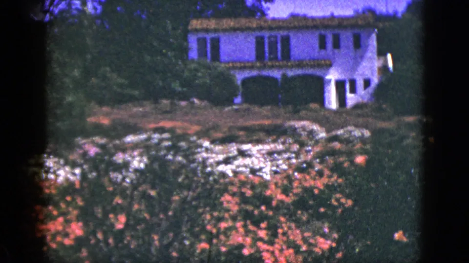 1953 Palm Springs home-movie frame showing a white stucco house with terracotta roof partly obscured by flowering garden plants.