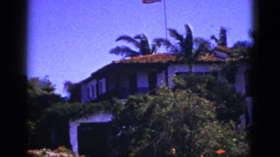 1953 Palm Springs exterior view of a stucco house with terracotta roof, palms, dense greenery, and a small American flag above.