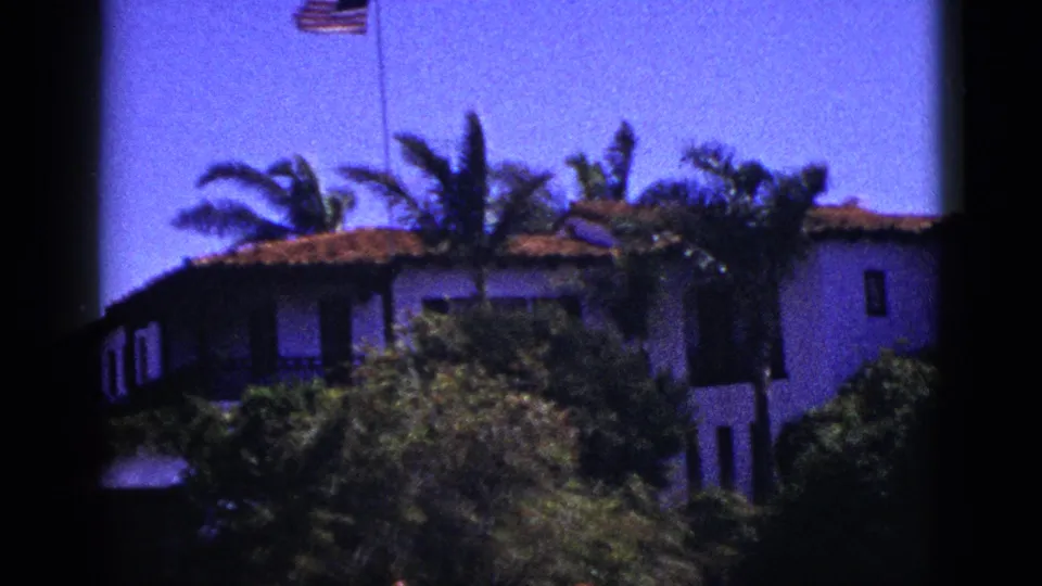 1953 Palm Springs frame with a tiled-roof stucco house partly hidden by palms and foreground shrubs under bright sky.
