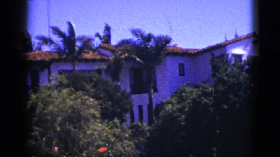 1953 Palm Springs home-movie view of a stucco house behind dense greenery, with palm fronds and terracotta roof visible.