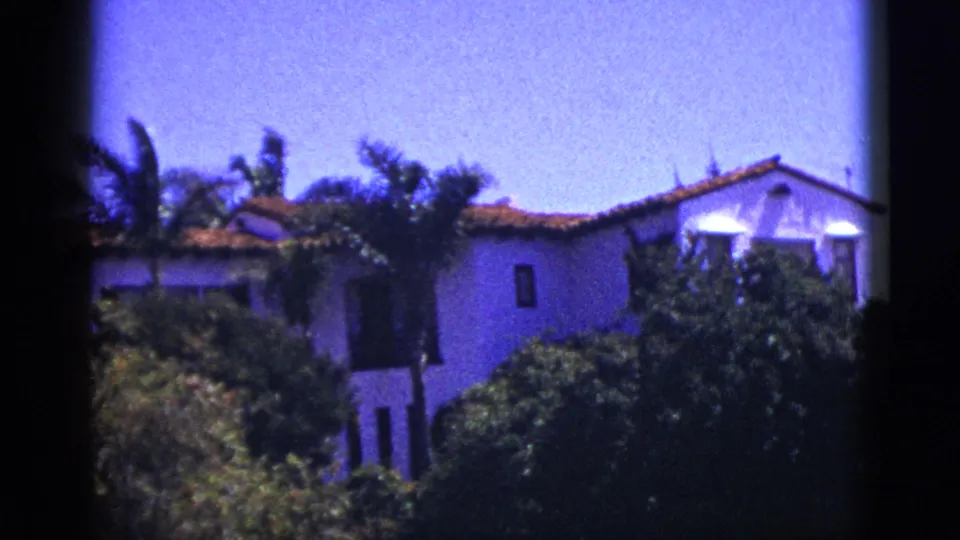 1953 Palm Springs 8mm frame showing a white hacienda-style building with red tile roof, palms, hedges, and dark frame edges.