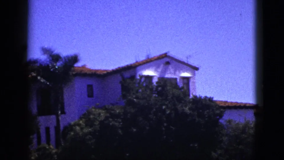 1953 Palm Springs exterior view of a white stucco-like building with terracotta roof, thick greenery, and a dark vertical frame in the foreground.