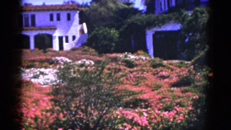 1953 Palm Springs home-movie frame of a stucco house, garage, flowering beds, and thick planted landscaping.