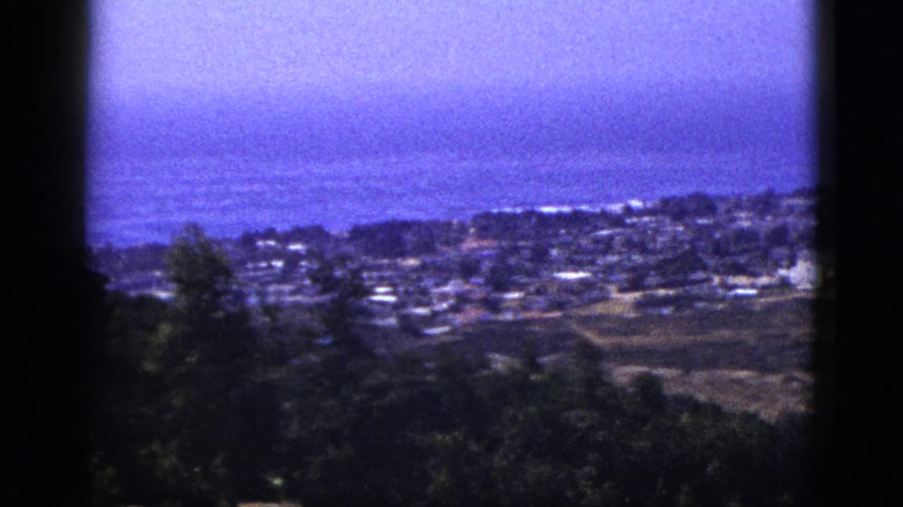 1953 8mm home-movie frame looking from a dark foreground hillside over clustered buildings toward a hazy ocean horizon.