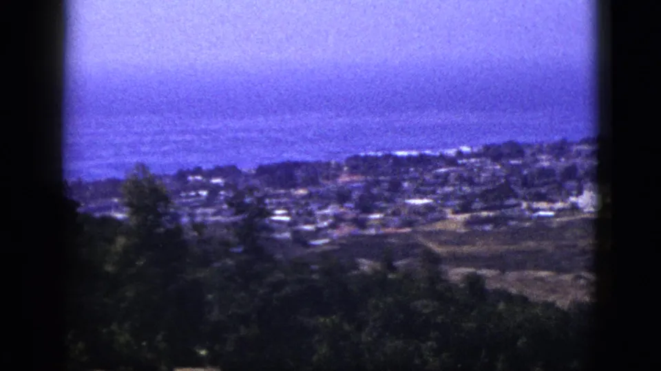 1953 8mm home-movie frame looking from a dark foreground hillside over clustered buildings toward a hazy ocean horizon.