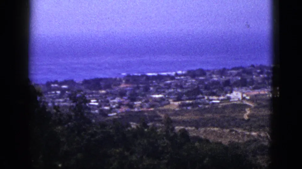 1953 8mm frame with a soft, grainy overlook across low buildings and open ground toward a blue-purple ocean horizon.