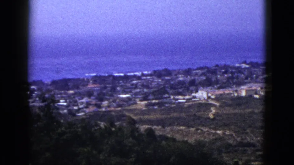 1953 8mm home-movie frame showing a distant town or neighborhood near an ocean horizon, framed by dark gate edges and shadowed foreground vegetation.