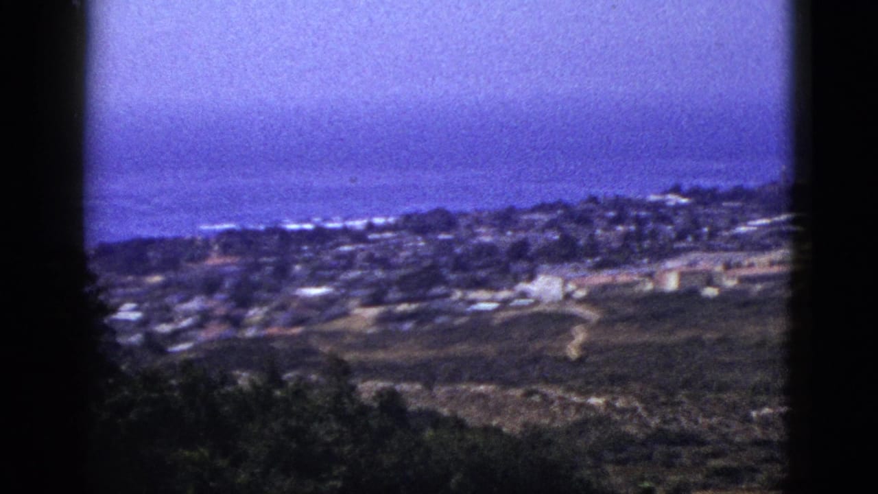 Distant town and hillside viewed through dark 8mm frame borders under a blue hazy sky.