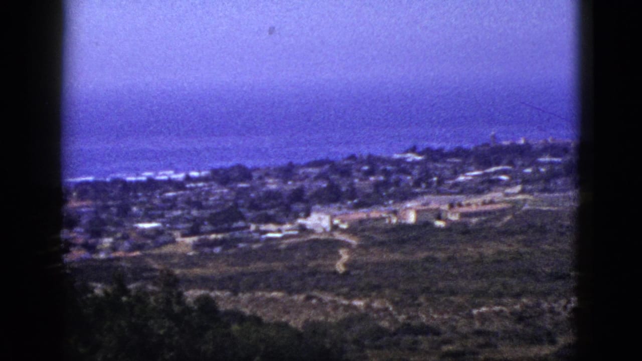 Panoramic hillside view of buildings, open land, and a blue horizon within dark film borders.
