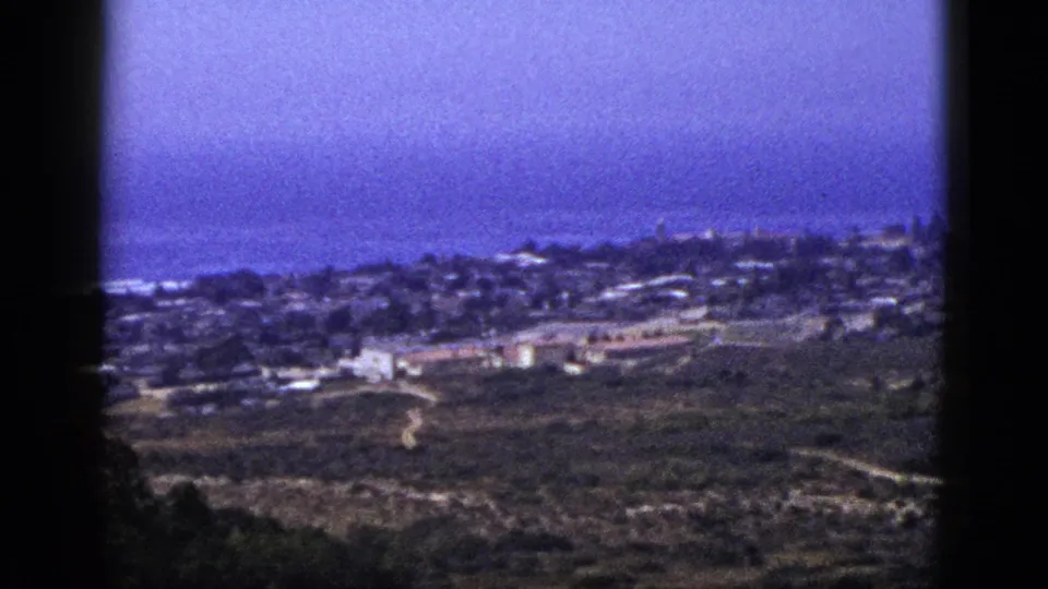 Mid-century 8mm landscape frame with a town edge, open fields, pale roads, and a blue distant horizon seen from above.