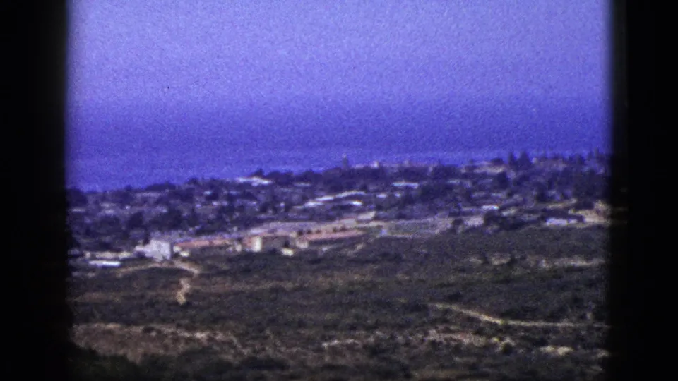 Vintage 8mm hillside panorama with distant low buildings, winding roads, open ground, and strong blue-purple film color cast.