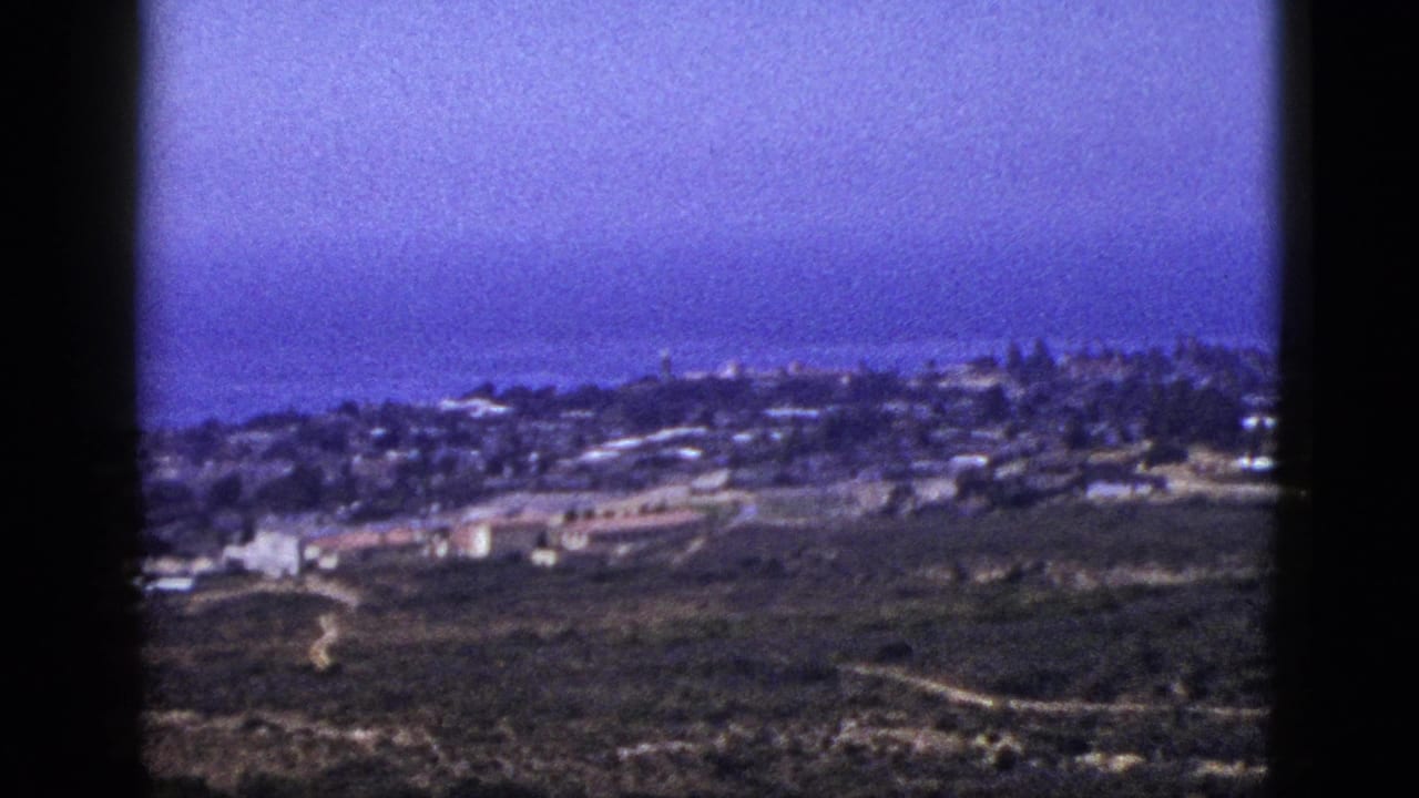 1953 California 8mm frame showing a coastal settlement below an elevated viewpoint, with open hillsides and the Pacific Ocean in the distance.